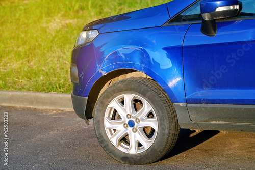 Fotografija Car with front end damage in parking lot, front fender with large dent and visible scratches on blue car body, impact and the need for repair