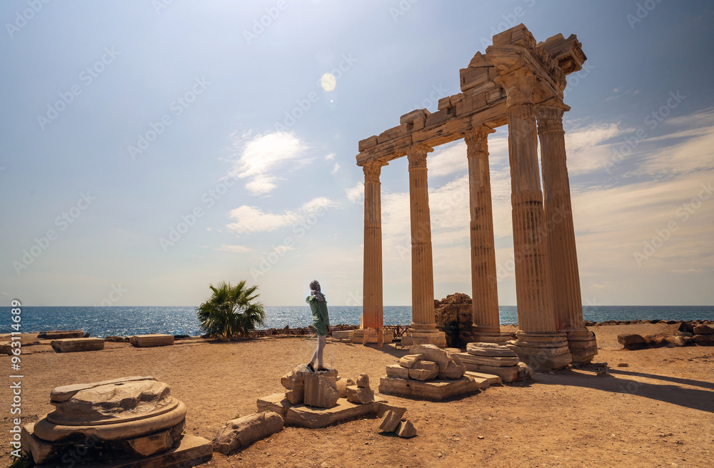 A woman in burka posing against breathtaking view of the Temple of Apollo ruins in Side, Turkey, with the Mediterranean Sea in the background.