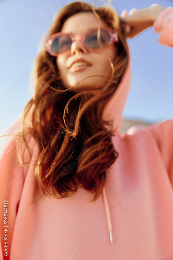 Stylish woman in pink hoodie and sunglasses striking a pose against vibrant blue sky background Stylish woman in pink hoodie and sunglasses striking a pose against vibrant blue sky background