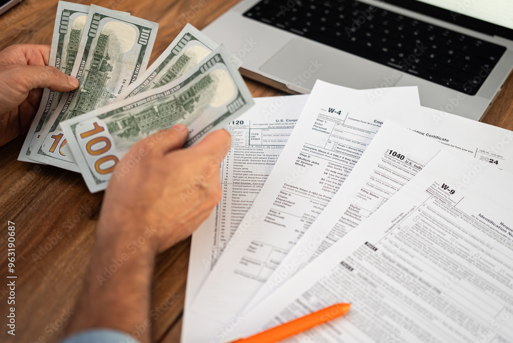 Man freelancer holds dollar bills in hands counting income for filling tax forms to report income meeting deadlines in accordance with strict laws at desk with laptop Man freelancer holds dollar bills in hands counting income for filling tax forms to report income meeting deadlines in accordance with strict laws at desk with laptop