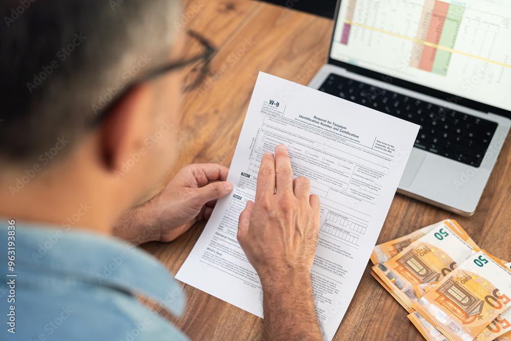 Naklejka premium Small business owner examines W-9 tax form to provide Taxpayer Identification Number for reporting income paid by clients at table alongside laptop and euro bills
