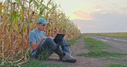Farmer sits on the edge of a dirt road working on a laptop with corn fields stretching out behind him.