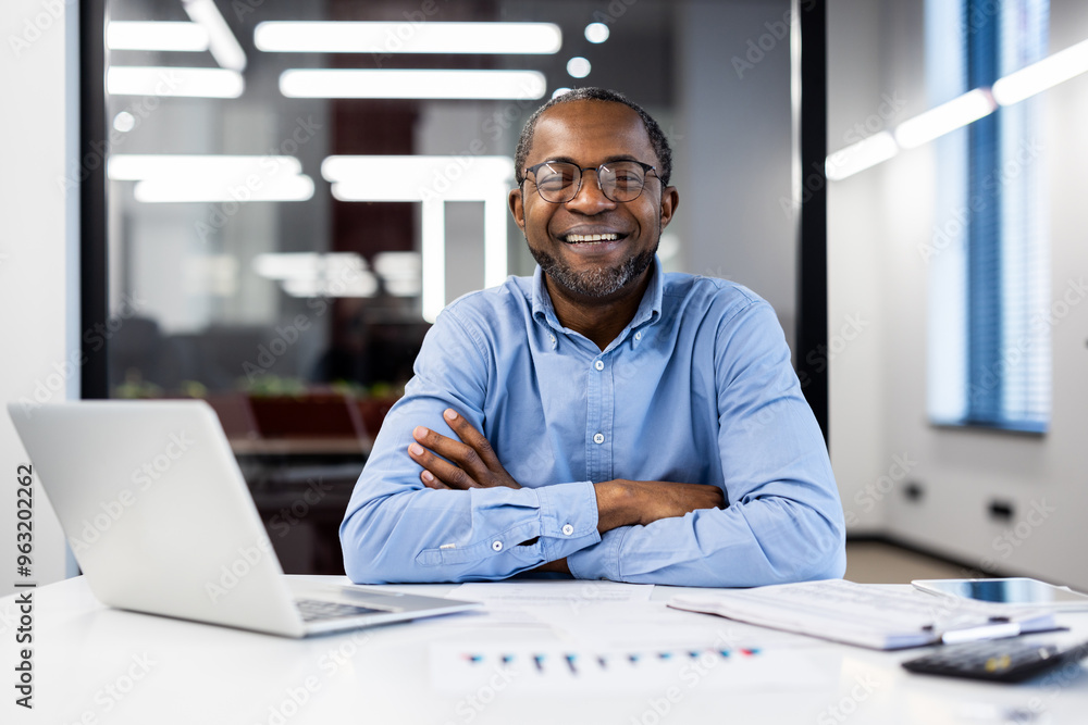 Obraz premium Businessman wearing glasses smiling confidently in office environment. He sits at desk with laptop and documents, exuding positivity and professionalism.