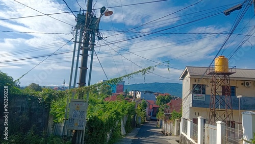 A narrow street in a residential area with power lines, houses, and a water tank.