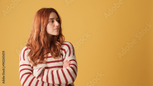 Red-haired girl dressed in sweater, looking away to show mockup space on yellow background in studio. Beautiful girl poses in studio with blank space on yellow wall for your promotion