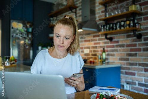 Young woman using smartphone in kitchen while working from home