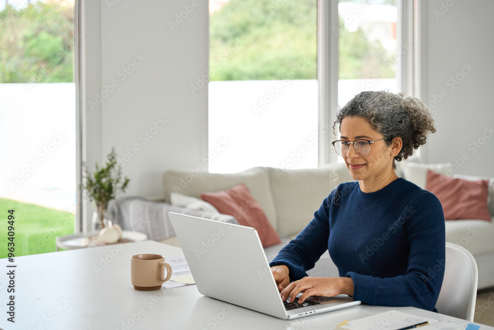 Happy middle aged woman sitting at table at home and using laptop in modern house. Mature 50 years old lady wearing glasses remote working online from home typing at computer in living room. Happy middle aged woman sitting at table at home and using laptop in modern house. Mature 50 years old lady wearing glasses remote working online from home typing at computer in living room.