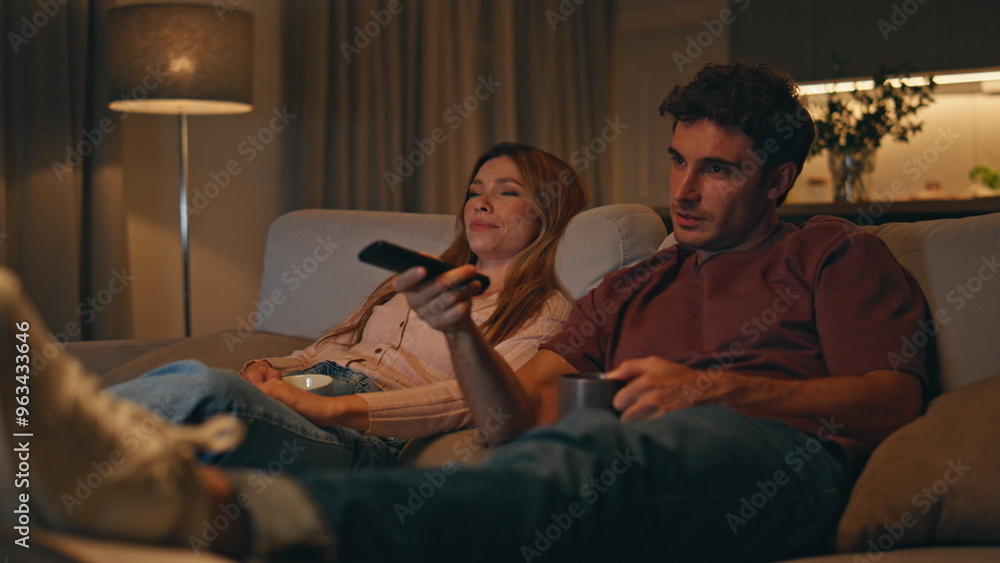 Fototapeta premium Smiling spouses watching tv sitting at cozy living room interior late evening