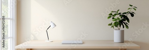 A clean and modern home office desk with a white lamp, a laptop, and a potted plant. The desk is made of natural wood, and the background is a light beige wall. The window provides a view of the outdo photo