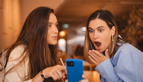 Portrait of excited impressed two women sisters friends fellows use devices recommend select choose decide discount options, point finger on phone, screaming wow, open mouth, omg, sitting in cafe.