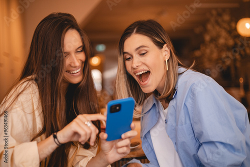 Portrait of excited impressed two  women sisters friends fellows use devices recommend select choose decide discount options, point finger on phone, screaming wow, open mouth, omg, sitting in cafe.