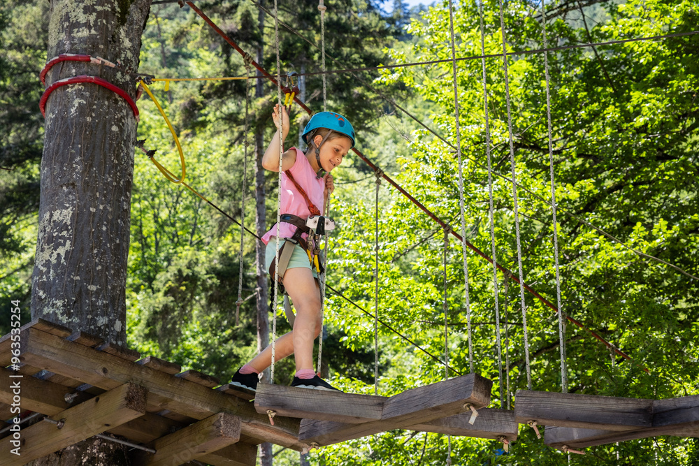 A girl in a pink shirt and blue helmet cautiously moves through a high ropes course in the woods, walking on a narrow wooden platform high above the ground, surrounded by lush green trees A girl in a pink shirt and blue helmet cautiously moves through a high ropes course in the woods, walking on a narrow wooden platform high above the ground, surrounded by lush green trees