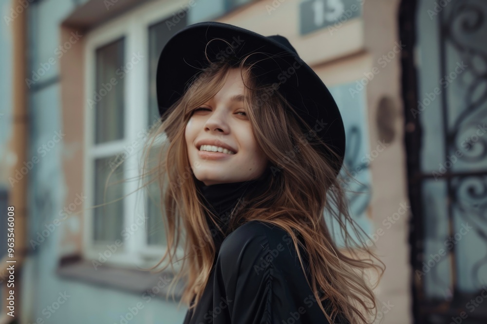 A woman wearing a black hat and a black dress is smiling. She is standing in front of a building with a window A woman wearing a black hat and a black dress is smiling. She is standing in front of a building with a window