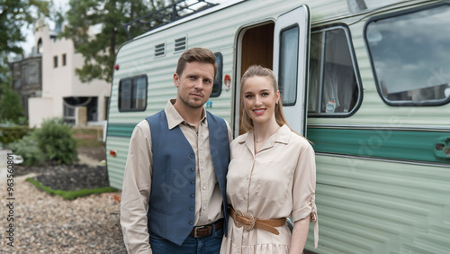 A couple poses together in front of a vintage camper, dressed in casual yet stylish clothing, evoking a sense of adventure and retro charm in an outdoor setting.