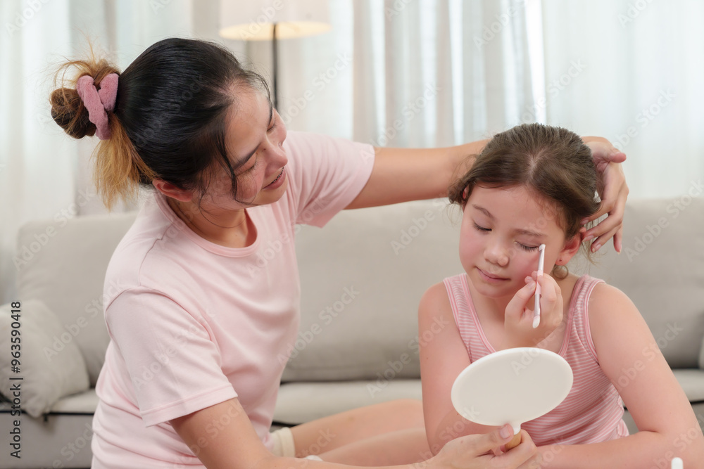 Fototapeta premium Asian mother and daughter sharing a joyful moment as they apply makeup together. Daughter holds a brush and mirror, while mother offers guidance. Surrounded by beauty products, love and happiness.