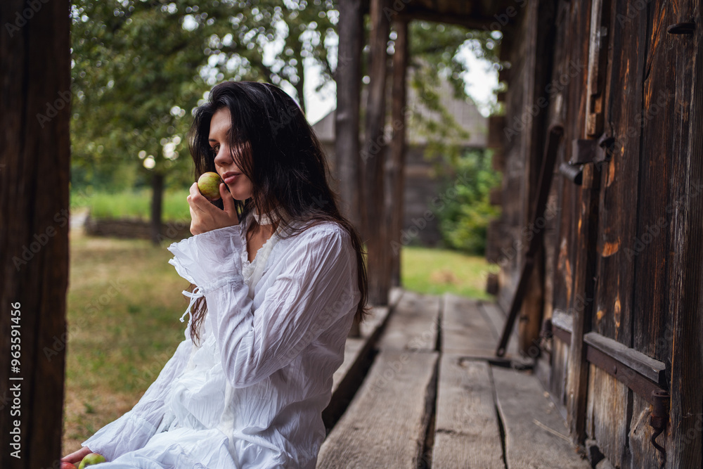 Retro style. Young beautiful sexy girl with black chic hair in a white dress sitting on wooden stairs near an old wooden house and eating an apple. Retro style. Young beautiful sexy girl with black chic hair in a white dress sitting on wooden stairs near an old wooden house and eating an apple.