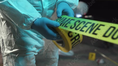 A forensic scientist rolling out police tape at a crime scene at night, while other experts collect evidence in the background.