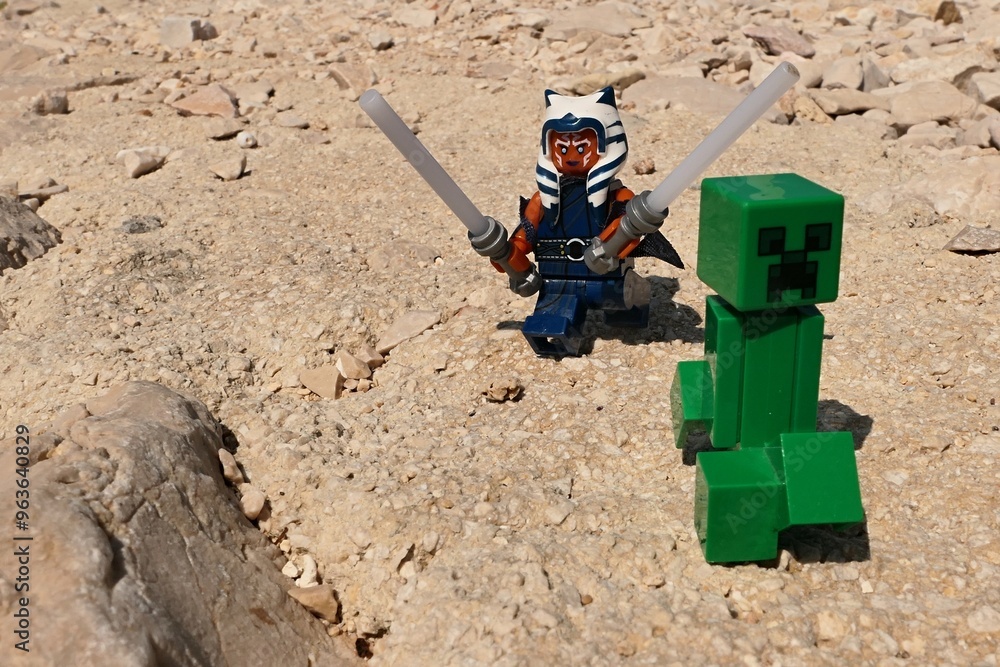 Naklejka premium LEGO Star Wars figure of female Togruta Jedi Ahsoka Tano with two lightsabers chasing green explosive LEGO Minecraft mob Creeper on shoreline rocks, summer daylight sunshine. 