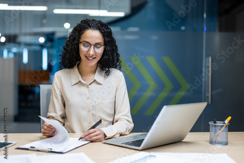 Confident Hispanic businesswoman analyzing documents at desk with laptop, demonstrating focus and professionalism in modern office environment.