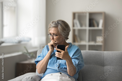 Foto Unhappy older woman sitting on couch, holding smartphone feels frustrated and co