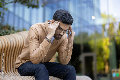 Man with glasses on outdoor bench holding head in hands. Depicts stress, contemplation, worry, or headache. Urban setting suggests work or personal struggles. Expression reveals tension, pondering