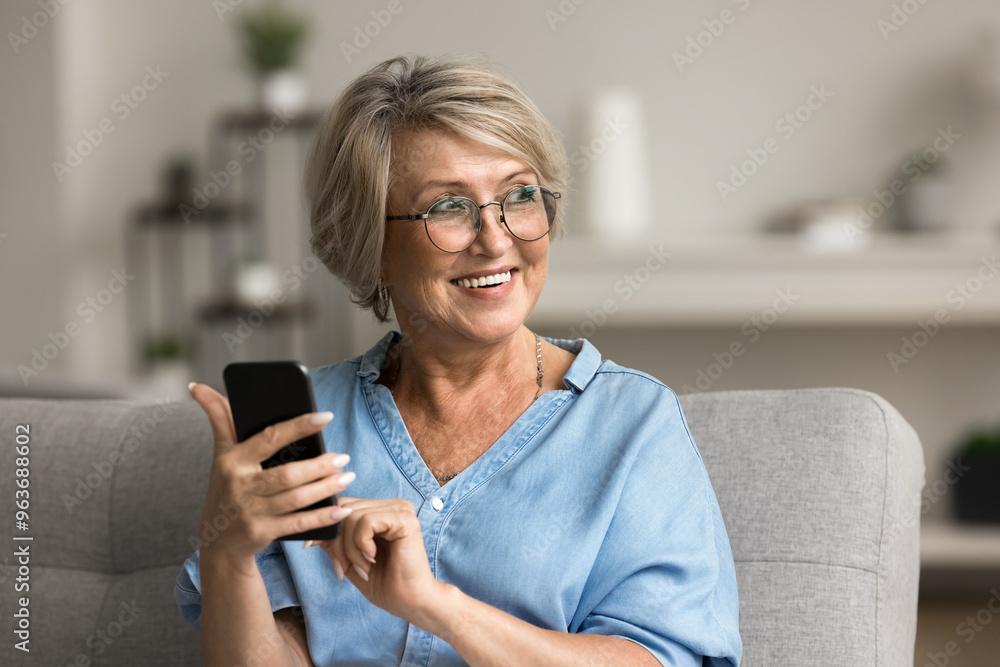 Pretty middle-aged woman in glasses looking away holding smartphone smile, resting on sofa at home, making call, browse wireless internet, search information, spend free time online on leisure indoors Pretty middle-aged woman in glasses looking away holding smartphone smile, resting on sofa at home, making call, browse wireless internet, search information, spend free time online on leisure indoors