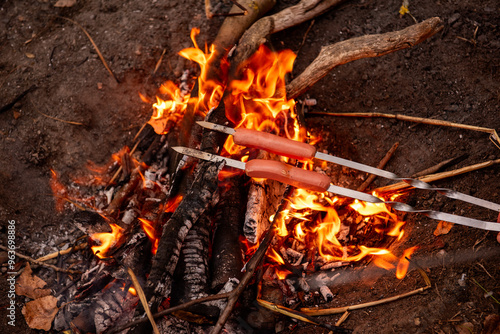 Two hot dogs sausages roasting on skewers over an open campfire with bright flames and burning logs in an outdoor setting. photo