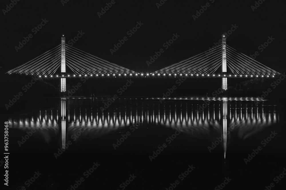 Fototapeta premium Side view of a suspender cables bridge over a river at night, with its reflection on the water. B&W image.
