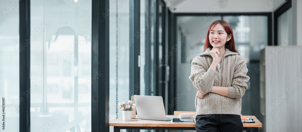 A young woman stands confidently in a modern office, with a laptop and documents on the desk, reflecting a professional work environment. A young woman stands confidently in a modern office, with a laptop and documents on the desk, reflecting a professional work environment.