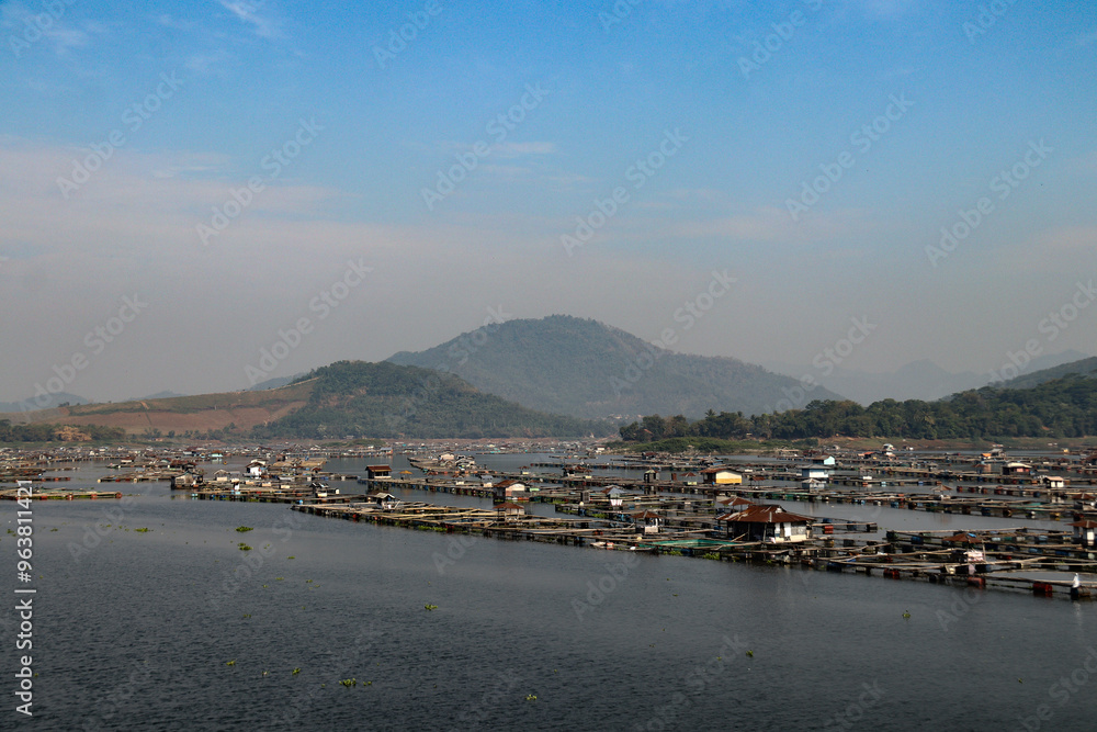 Fototapeta premium A stunning waterscape featuring fish farms floating on a lake, framed by distant green hills and mountains under a bright blue sky. A peaceful rural aquatic landscape