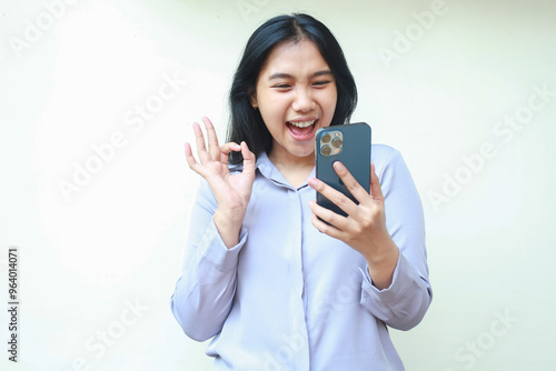 excited carefree asin young business woman holding mobile phone and gesturing ok sign while video calling, smiling female with approval pose wearing formal shirt isolated in white background photo