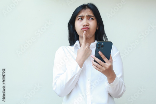 elegant asian young business woman daydreaming using smartphone wearing white formal shirt with finger on chin looking up confused what to buy in online shopping sale standing over isolated background