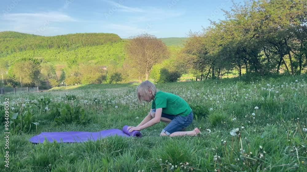 Young boy wearing a green t shirt is rolling a purple yoga mat in a field with trees and hills in the background Young boy wearing a green t shirt is rolling a purple yoga mat in a field with trees and hills in the background