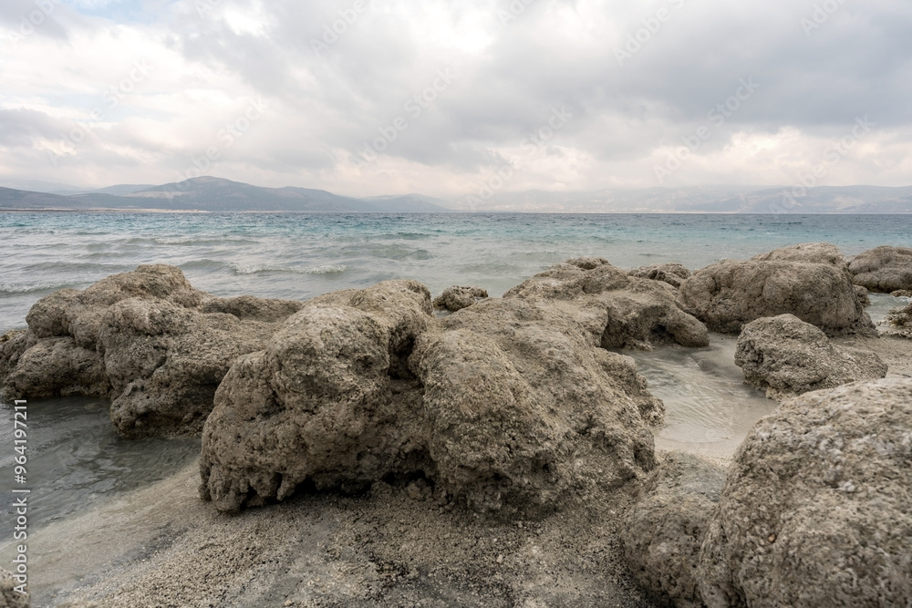 Turquoise waters and white mineral beaches of Lake Salda, Turkey, with a cloudy sky and rocky formations in the background.
