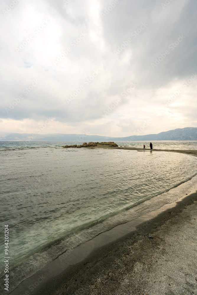 A view of Lake Salda, Turkey, showcases a crescent-shaped bay with pristine white sand and clear turquoise waters. Dark storm clouds loom overhead, adding a dramatic touch to the scene.