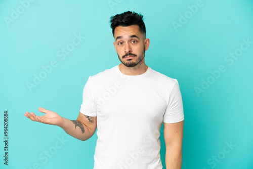 Young caucasian man isolated on blue background making doubts gesture