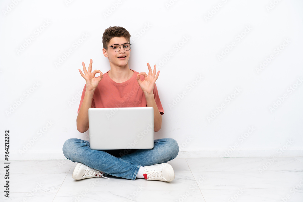 Teenager Russian man holding pc sitting on the floor isolated on white background showing an ok sign with fingers Teenager Russian man holding pc sitting on the floor isolated on white background showing an ok sign with fingers