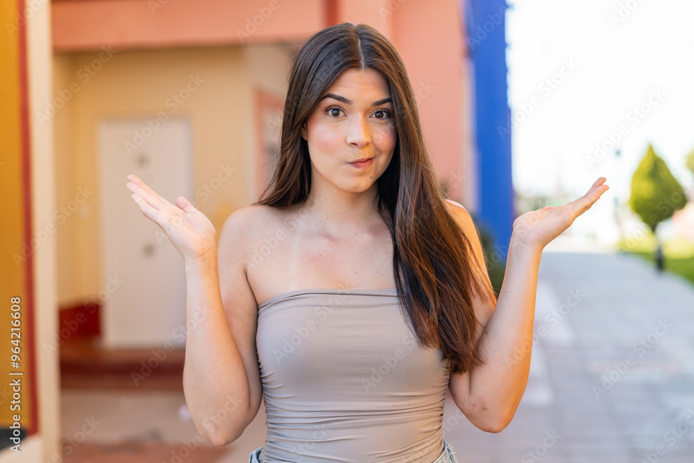Young pretty Brazilian woman at outdoors having doubts while raising hands Young pretty Brazilian woman at outdoors having doubts while raising hands