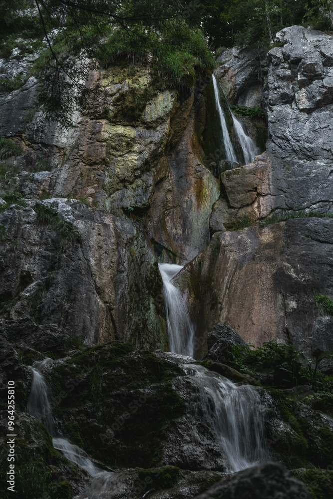 A multi-tiered waterfall cascades over rugged, moss-covered rocks in a forested area. The water flows gracefully through the steep rock formations, creating a serene and picturesque natural scene. A multi-tiered waterfall cascades over rugged, moss-covered rocks in a forested area. The water flows gracefully through the steep rock formations, creating a serene and picturesque natural scene.