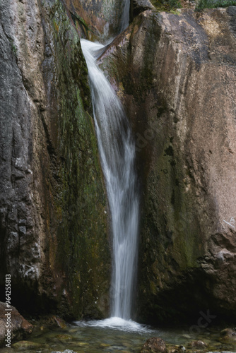 A narrow waterfall gracefully flows down between moss-covered rocks into a clear pool below. The contrast between the soft water and rugged rocks creates a serene and peaceful natural scene. 