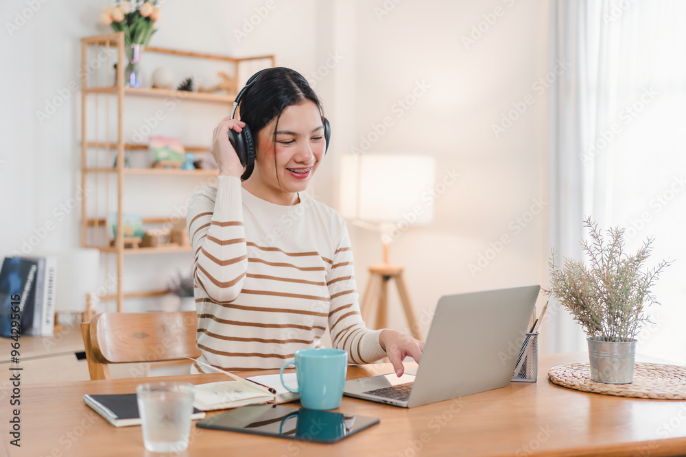 Smiling Asian woman relaxing on a cozy desk, wearing headphones, using a laptop, enjoying a comfortable home.