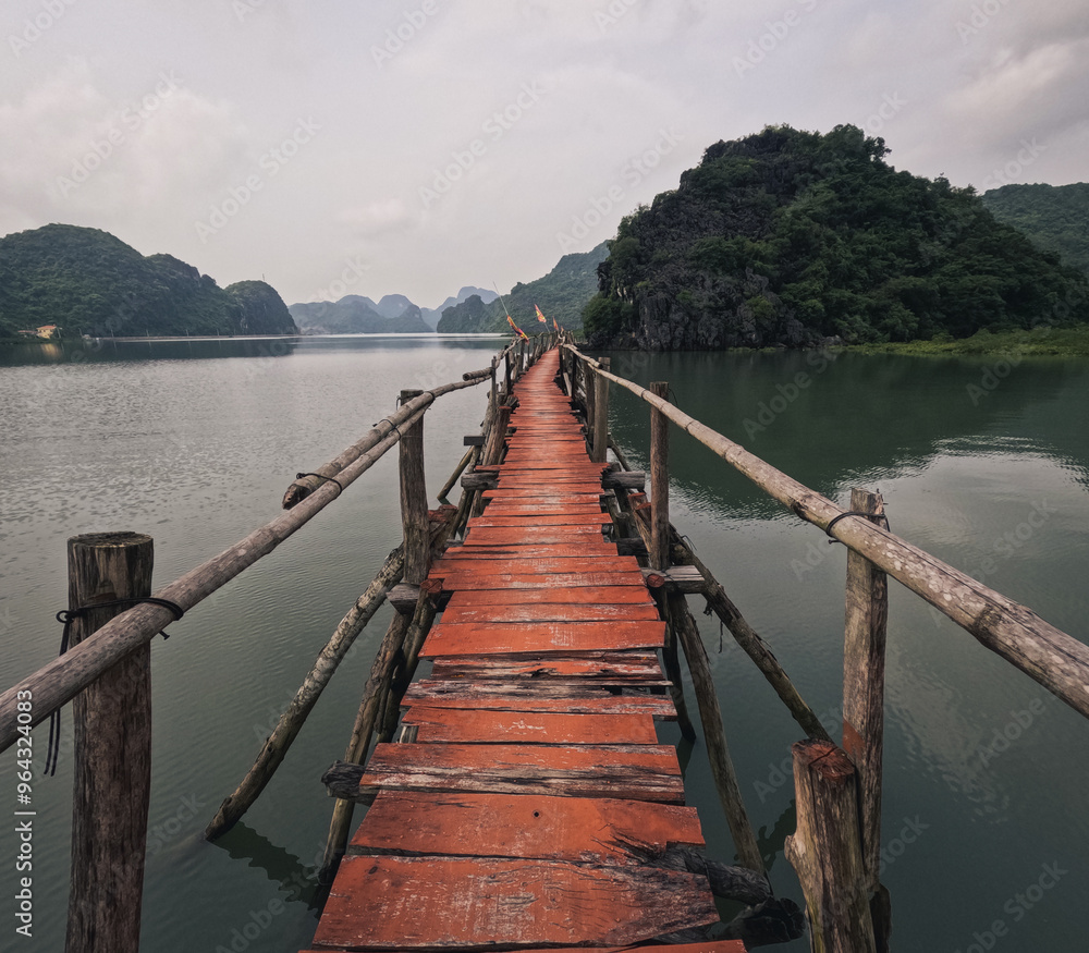 Naklejka premium Red wooden bridge and boardwalk over emerald waters in Cat Ba Island, Vietnam, framed by dramatic karst limestone mountains and tropical landscape.