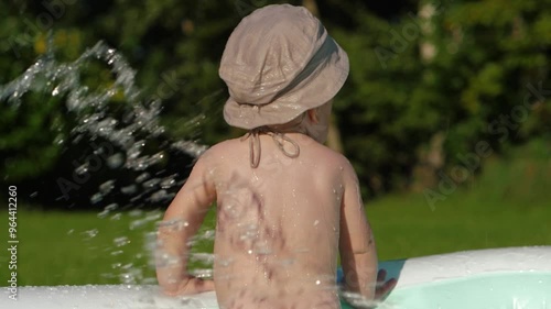 Infant child in stylish sun panama hat standing backside in inflatable pool, embodying summer and creating unforgettable memories. Baby boy stands with his back, splashing in backyard, slow motion