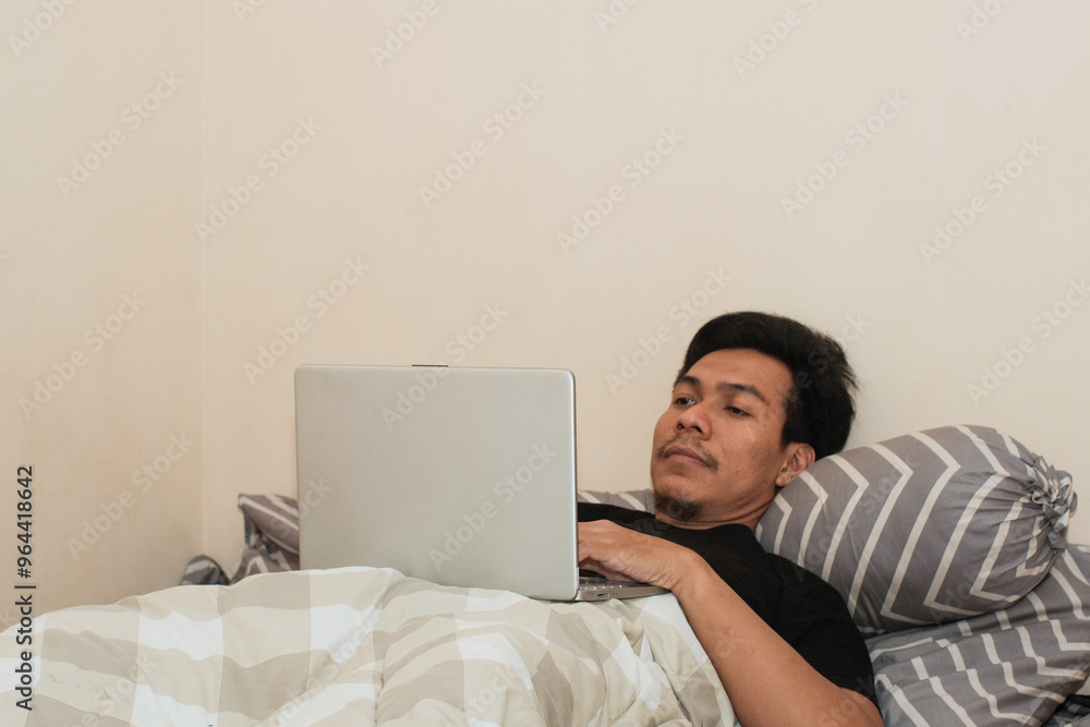Always online. A Southeast Asian man working on laptop in bed, copy space. Early morning. Pleasant awakenin Always online. A Southeast Asian man working on laptop in bed, copy space. Early morning. Pleasant awakenin
