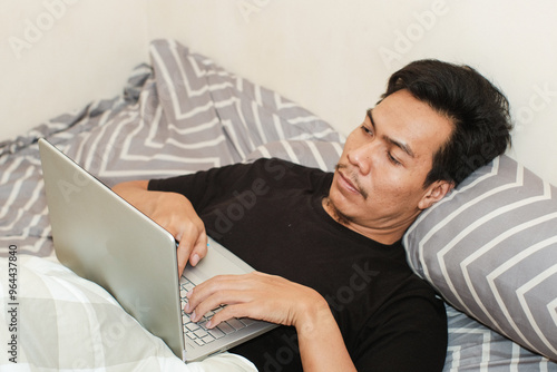 Always online. A Southeast Asian man working on laptop in bed, copy space. Early morning. Pleasant awakenin photo