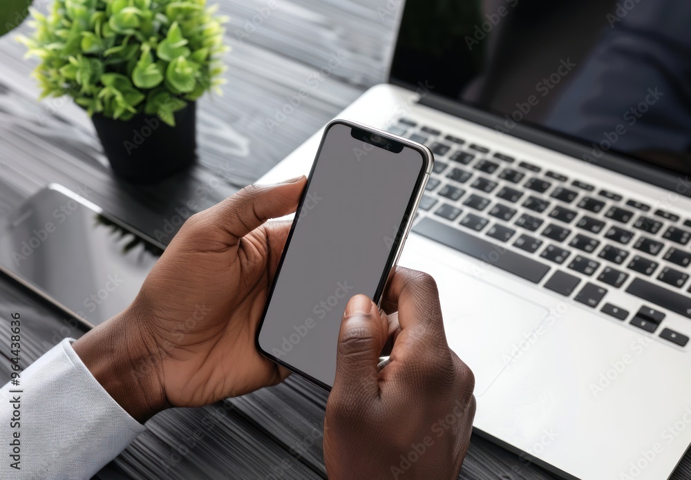 Hands holding a smartphone with a blank screen next to a laptop on a desk. Modern technology and digital communication concept. Hands holding a smartphone with a blank screen next to a laptop on a desk. Modern technology and digital communication concept.
