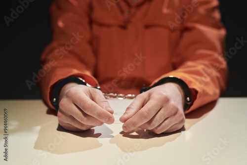 Person's hands handcuffed on table in dimly lit room, wearing orange jumpsuit suggesting incarceration. Shadows add intense, moody atmosphere reflecting seriousness of situation