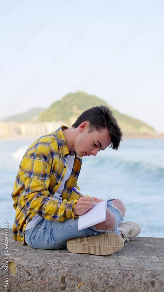 Young man writing on a notebook outdoors. He is sitting outdoors, at the seaside. Young man writing on a notebook outdoors. He is sitting outdoors, at the seaside.