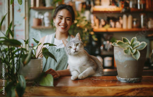 A short-haired cat sitting on a wooden coffee bar counter among potplants with a woman barista or waitress wearing an apron.   photo