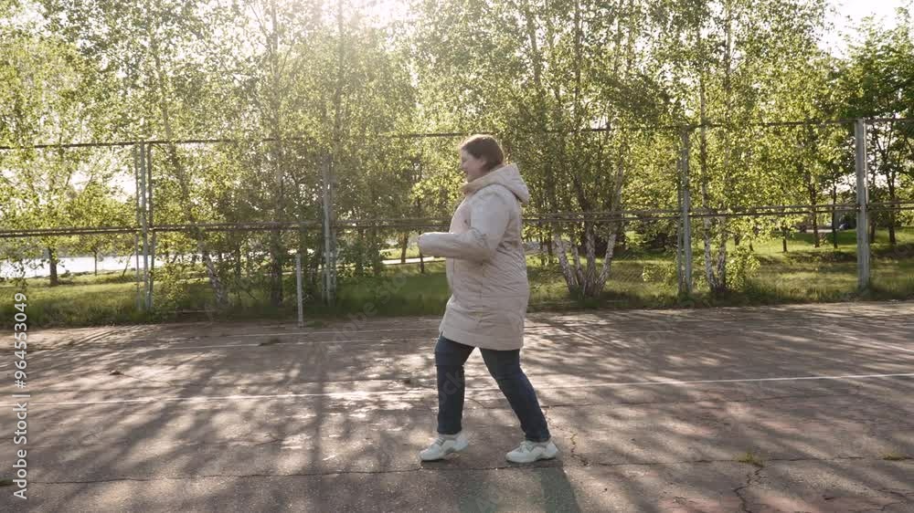 Plus size woman in jacket jumping cheerfully on old sports ground in park, side view. Middle aged nordic female person with smile exercising and warming up on sunny day in early spring, wide view. Plus size woman in jacket jumping cheerfully on old sports ground in park, side view. Middle aged nordic female person with smile exercising and warming up on sunny day in early spring, wide view.